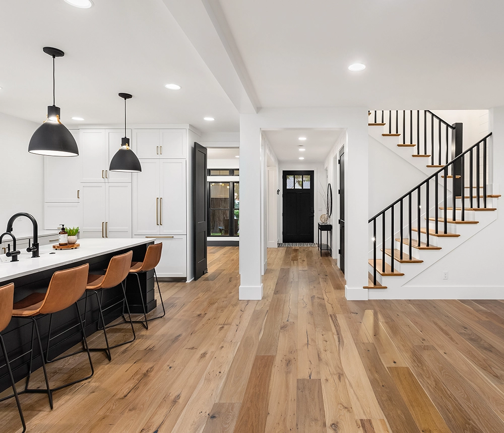 A wood flooring in an open-space kitchen with white cabinets, large kitchen island with leather stools, and black pendant lights