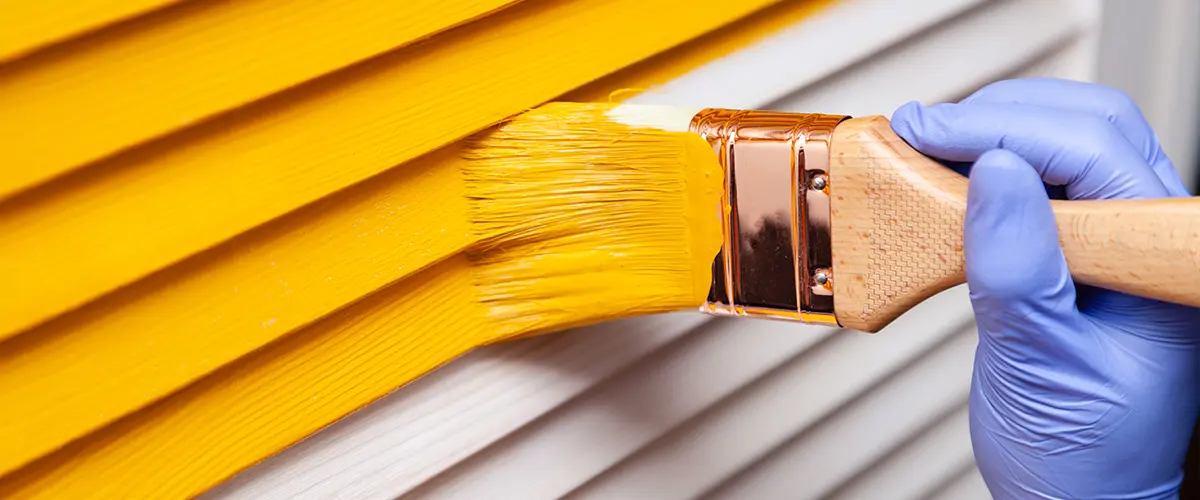 Close-up of hand wearing blue glove painting a wooden surface yellow with a brush, showcasing exterior painting in Reno.