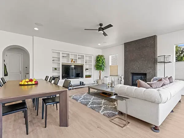 Cozy living room with a vaulted ceiling, large windows, and a white brick fireplace, featuring beige armchairs, a gray sofa, and a patterned area rug on dark hardwood flooring.